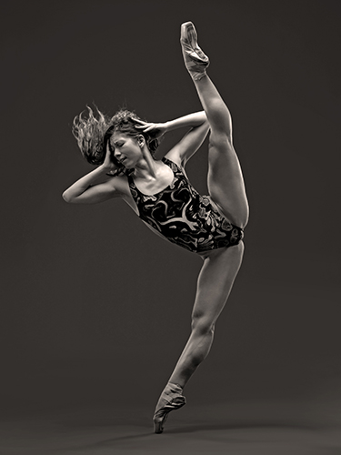 A ballerina in a geometrically patterned leotard gracefully lifts one leg straight up above her head while standing on the point of her toes. She has an emotional expression while both hands touch her head.