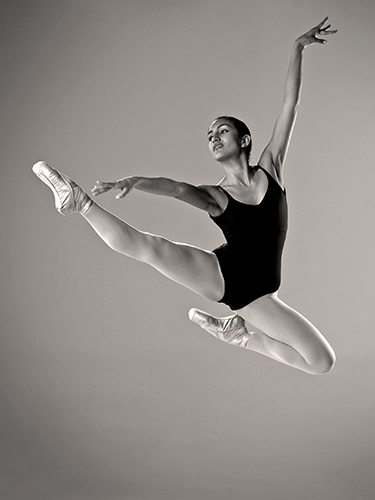 A ballet dancer in mid-air performs a grand jeté. She wears a black leotard, white tights, and pointe shoes. Her left arm is extended upward, her right arm outward, and her gaze is focused straight ahead.