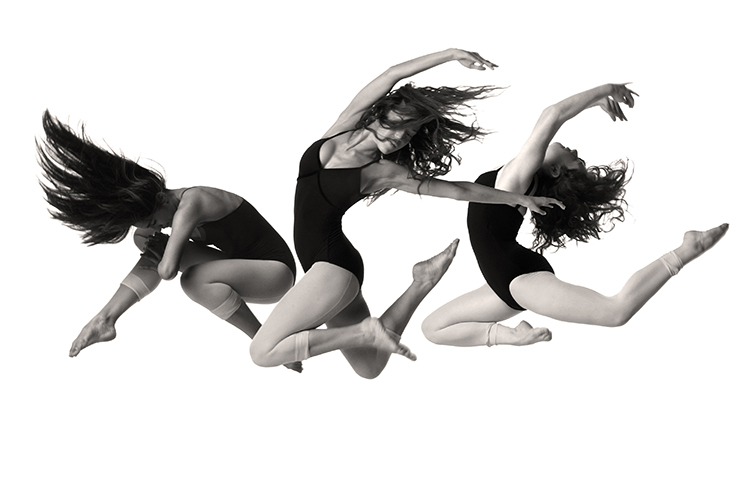 Three dancers in black leotards gracefully leap in mid-air against a white background, showcasing dynamic movement and flowing hair. The choreography highlights athleticism and elegance.