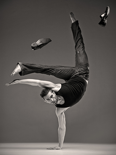 A man performs a one-handed handstand, wearing jeans and a black tank top. His shoes are airborne, and his expression is focused and intense.