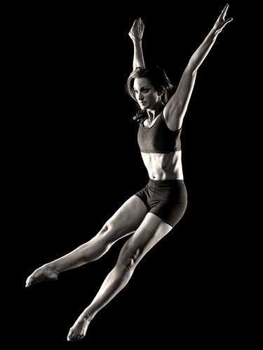 A woman in mid-air against a dark background, wearing a sports bra and shorts. Her arms are raised, and she exhibits a graceful, athletic pose. The lighting highlights her toned muscles.