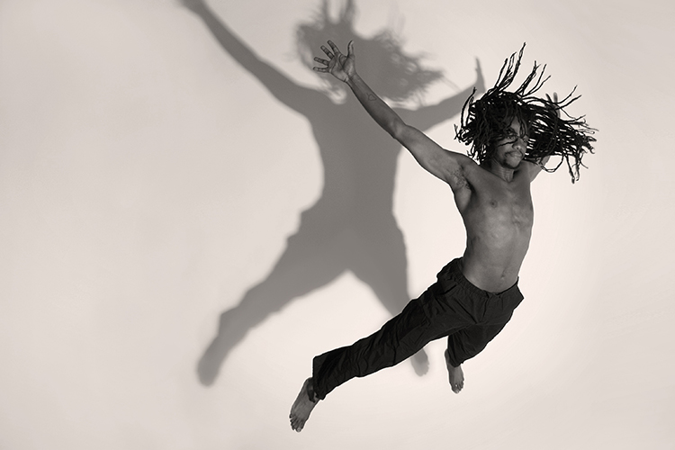 A man with long dreadlocks is leaping gracefully in mid-air against a plain background. His shadow projected behind him adds a dramatic effect. He is wearing dark pants and is shirtless, with arms and legs extended in a dynamic pose.