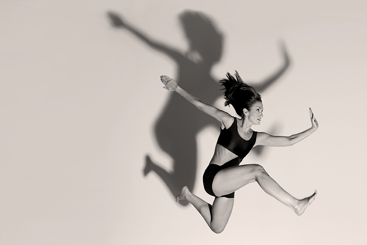 A dancer in mid-air against a light background, casting a dramatic shadow. She is wearing a two-piece dance outfit, and her hair is floating up. Her pose is expressive and dynamic, emphasizing movement and grace.