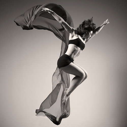 A dancer in a black sports bra and shorts is leaping beside a piece of shear fabric floating in the air. Her knees are bent up together and her arms are up and out. 