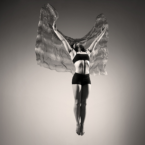 A dancer captured mid-leap, wearing a dark outfit and surrounded by flowing fabric. The grayscale image highlights her athleticism and grace against a simple studio backdrop.