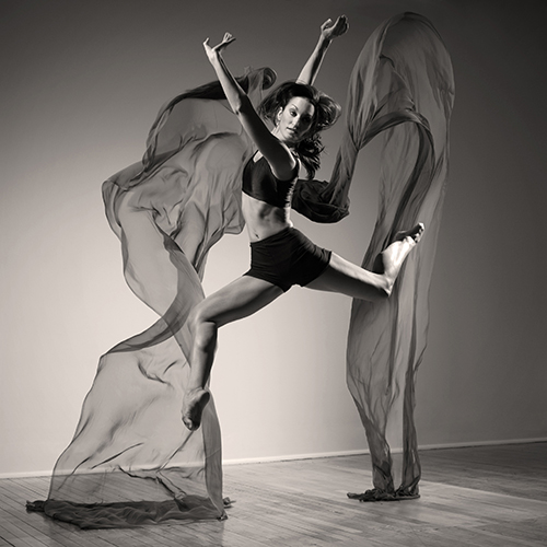 A dancer jumps gracefully in mid-air, arms reaching upward, with flowing fabric resembling wings. She wears athletic shorts and a crop top. The monochrome background adds a dramatic effect, highlighting the movement and elegance.