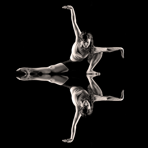 A dancer in a dynamic pose is reflected on a glossy black surface, creating a symmetrical effect. Her right leg is extended while her left leg is bent, and her arms are gracefully positioned. The background and reflection are dark, highlighting her form.