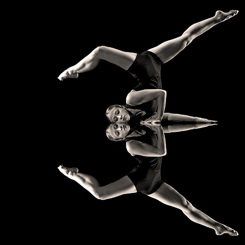 A dancer in a black outfit performs an acrobatic pose against a black background. The image is mirrored vertically, creating a symmetrical reflection. Her legs are extended in a split, and she holds a prayer position with her hands.