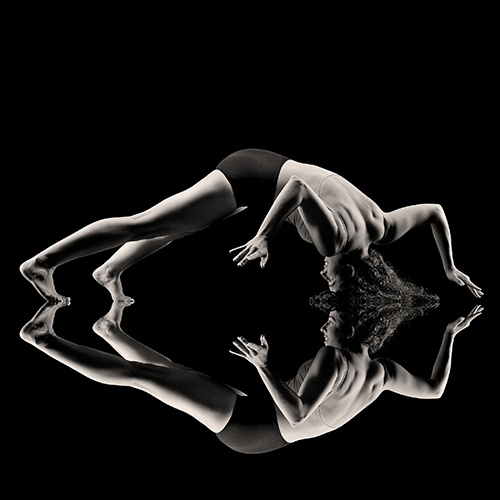 A woman performs an intricate yoga pose against a black background, with their body mirrored on the surface beneath them. The pose highlights strength and flexibility, creating a symmetrical visual effect.