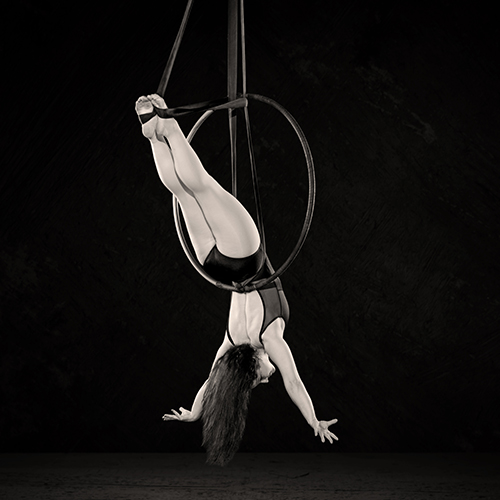 A woman performs an acrobatic pose on an aerial hoop, hanging upside down with legs extended and arms outstretched. The monochromatic image highlights the elegance and strength of the performer against a dark background.