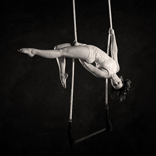 A person wearing a sparkling outfit performs an acrobatic pose on a trapeze. They hang upside down, holding the ropes with one hand and foot, against a dark background.