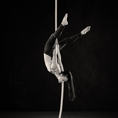 A woman performs an aerial acrobatics routine on a rope, gracefully hanging upside down with legs extended. The background is dark, emphasizing their balance and athletic skill.