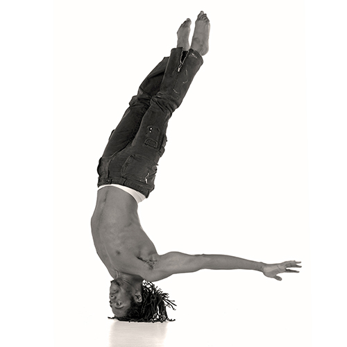 A man with dreadlocks is performing an impressive headstand with arms outstretched. They are shirtless and wearing jeans, showcasing strength and balance against a plain white background.