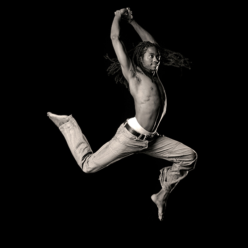 A man with long dreadlocks leaps gracefully against a black background. He is shirtless and wears jeans, with his arms raised and feet pointed, capturing a dynamic moment in mid-air.