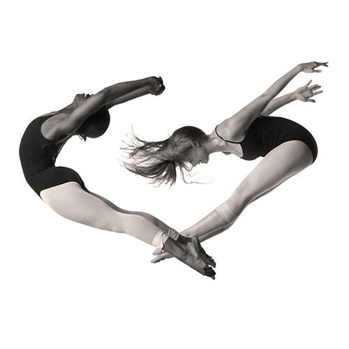 Two dancers in mid-air, forming a heart shape with their bodies. They wear black leotards and tights against a white background. One dancer arches backward, while the other extends forward, their feet touching at the center.