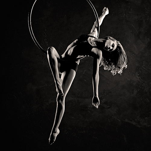 A black-and-white photo of a woman gracefully performing on an aerial hoop. She is wearing dancewear and her pose demonstrates strength and flexibility. The background is dark, emphasizing her form.