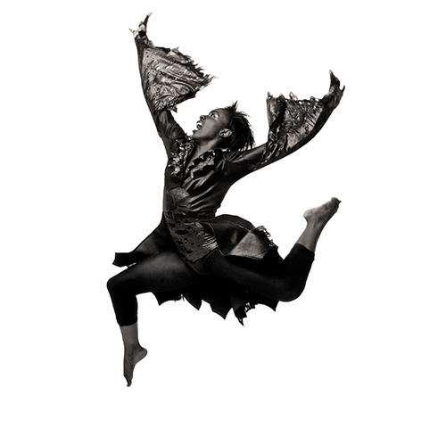 A dancer captured mid-air in a dynamic pose, wearing a textured costume resembling torn wings and a flowing dress. The image is in black and white, emphasizing movement and expression against a white background.