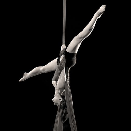 A woman performs an aerial silk routine, suspended upside down with one leg extended vertically and the other bent. They are holding the silk fabric with their arms, set against a dark background.