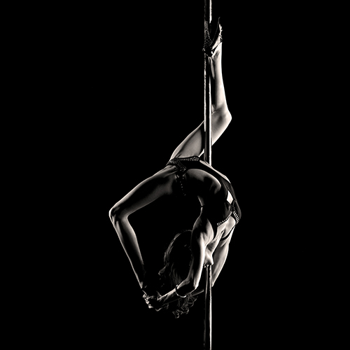 A woman performs an acrobatic pose on a pole in a silhouette against a dark background, showcasing strength and flexibility, with legs bent and arms reaching back.