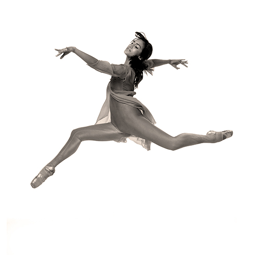 A dancer in mid-air performs a grand jeté, wearing a form-fitting bodysuit with a shear flowing top and pointe shoes, against a white background. The image captures the elegance and athleticism of the pose.