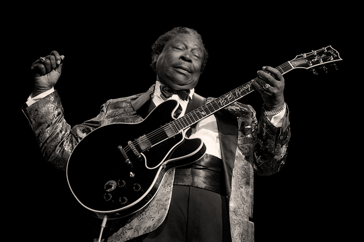 BB King wearing a patterned jacket plays an electric guitar passionately against a dark background.