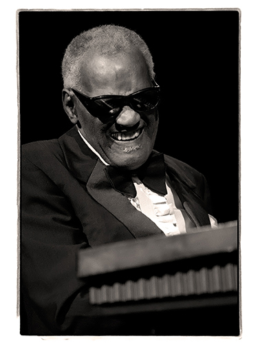 A black and white photo of Ray Charles wearing sunglasses and a tuxedo, smiling as he plays a keyboard. His expression is joyful and engaged in the music.