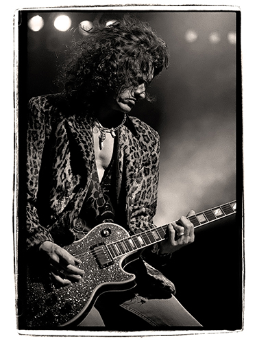 Joe Perry plays an electric guitar on stage, wearing a leopard print jacket. The background is dimly lit with spotlights visible, creating a dramatic atmosphere.