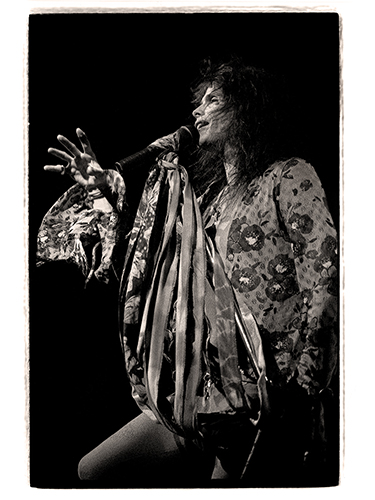 Steven Tyler performs passionately on stage, holding a microphone stand wrapped with flowing ribbons. He is wearing a floral-patterned outfit, captured in a vintage-style, black-and-white photograph.