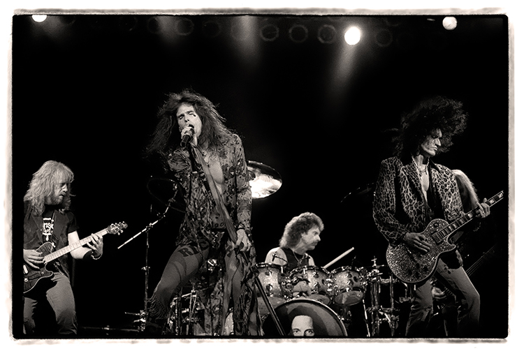 A black-and-white photo of a rock band Aerosmith performing on stage. The lead singer stands at the center with a microphone, surrounded by two guitarists. A drummer plays in the background. 