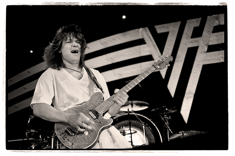 Guitarist Eddie Van Halen energetically plays on stage, wearing a light-colored outfit. Behind him, a logo with stylized letters is visible. The scene is lit dramatically, capturing the intensity of the performance.