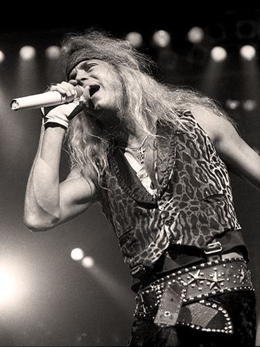 Bret Michaels passionately sings into a microphone on stage. He wears a patterned vest, a headband, and gloves. The background features stage lights, creating a dynamic concert atmosphere.