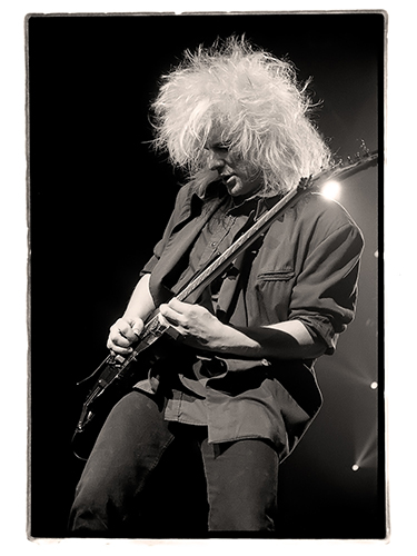 Black and white photo of CC DeVille playing an electric guitar on stage. The musician is wearing a dark jacket and pants sporting wild, voluminous hair. The background is lit by stage lights.