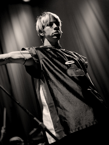 Anthony Kiedis performs on stage, holding a microphone stand. He is wearing a sleeveless shirt with a logo. The stage is dimly lit, creating a dramatic atmosphere with shadows.