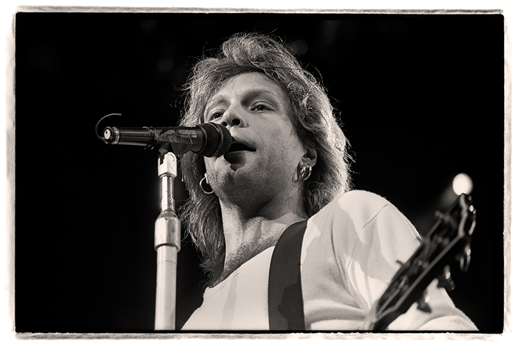 A black and white photo of Jon Bon Jovi singing into a microphone while holding a guitar. The stage lights are softly illuminating the background.