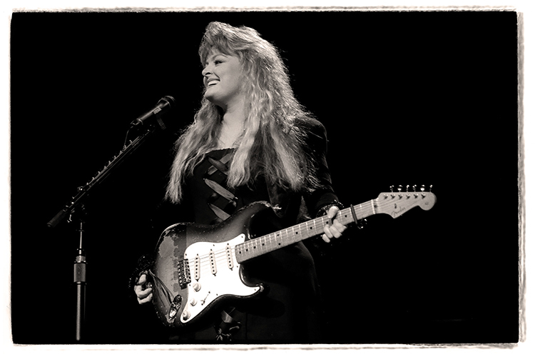 Black and white photo of Wynonna Judd holding an electric guitar on stage. She is smiling and wearing a stylish outfit. A microphone is positioned in front of her