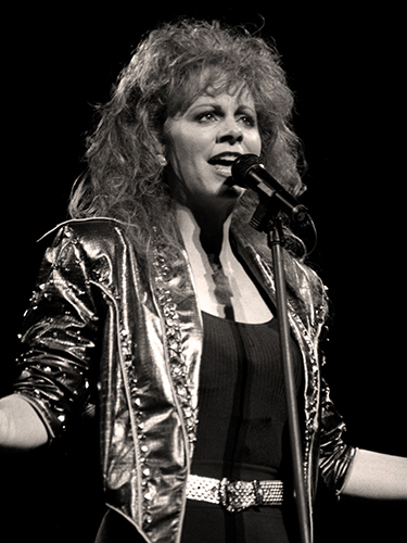 Reba McEntire singing passionately into a microphone on stage. She wears a shiny, studded jacket and has voluminous hair.