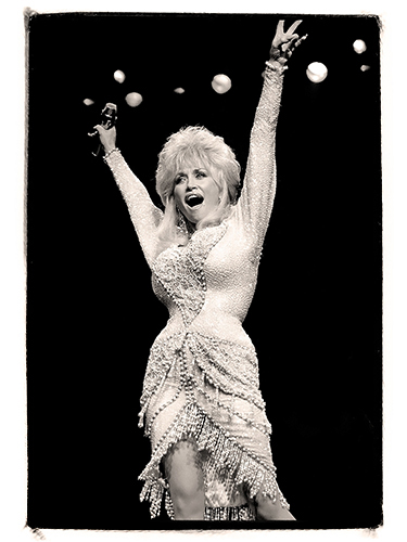 Dolly Parton wearing a sequined dress is on stage with a microphone in hand. She has her arms raised and is smiling, with stage lights visible in the background. The image is in black and white