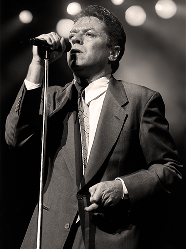 A black and white photo of Robert Palmer in a suit passionately singing into a microphone on stage. The background is blurred with circular lights, creating a dramatic atmosphere.