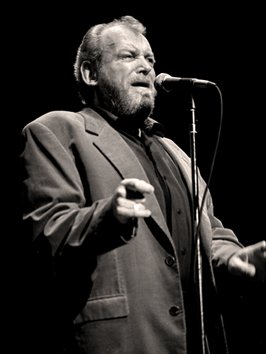 Joe Cocker, wearing a suit and shirt, sings into a microphone on stage. His expression is intense and passionate, and the background is dark, highlighting his presence under the spotlight.