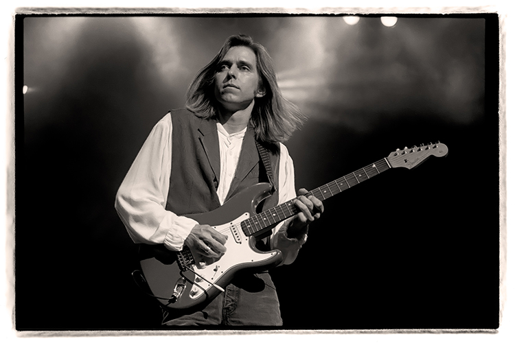 A black and white image of Eric Johnson playing an electric guitar on stage, with dramatic lighting and gentle smoke effects in the background. The musician is wearing a vest over a button-down shirt.