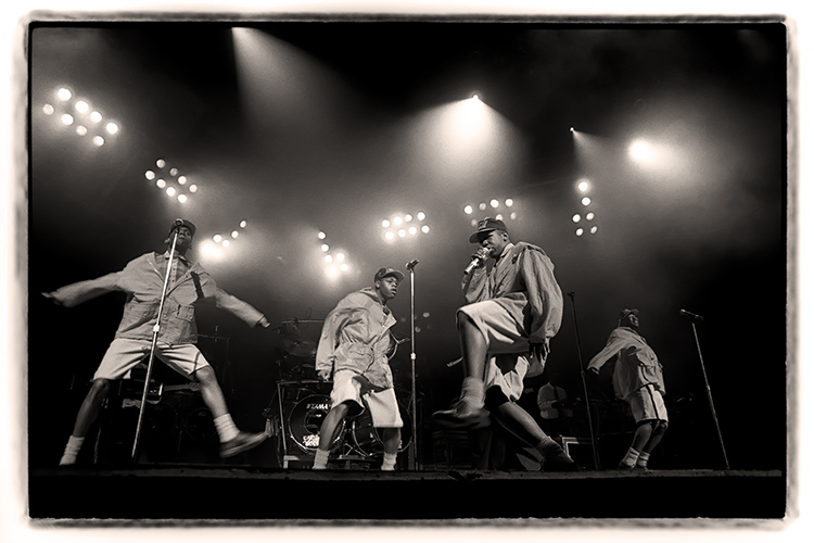 A black and white photo of Boys 2 Men performing energetically on stage. The four members dressed in matching outfits dance under bright stage lights, with microphones in hand. 