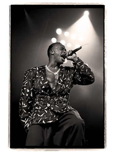 Black and white image of MC Hammer performing on stage, holding a microphone. He is wearing a patterned jacket and visible stage lighting creates a dramatic effect behind him.