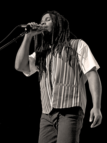 Ziggy Marley sings into a microphone, wearing a striped vest over a white shirt and pants, set against a dark background.