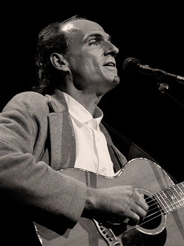 James Taylor, wearing a light-colored shirt and jacket, plays an acoustic guitar and sings into a microphone on stage. The image is in black and white.