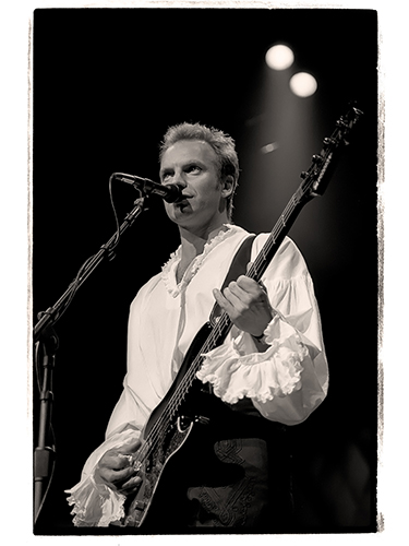 Sting playing a bass guitar and singing into a microphone on stage. He is wearing a frilly, long-sleeved shirt, lit by stage lights against a dark background.