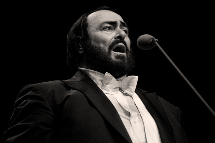 Opera tenor Luciano Pavarotti avidly singing into a microphone, wearing a tuxedo with a bow tie. The background is dark, emphasizing his expressive performance.