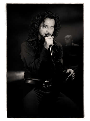 Dave Gahan from Depeche Mode sings subtlety into a microphone kneeling at the edge of the stage, dressed in a dark outfit. The black-and-white photo adds a dramatic and classic atmosphere