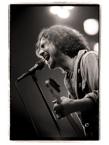 Black and white photo of Eddie Vedder from Pearl Jam passionately singing into a microphone while playing an electric guitar. The background is blurred with bright lights.