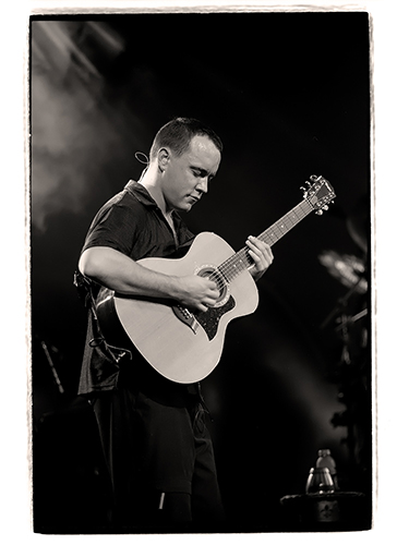 Dave Matthews playing an acoustic guitar on stage, captured in black and white. The scene has a spotlight and a foggy atmosphere