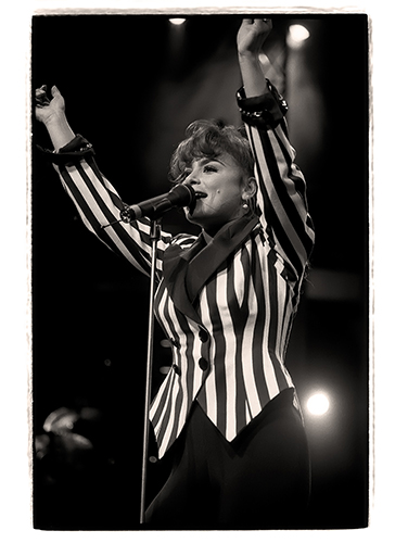 Paula Abdul performs energetically on stage, wearing a striped jacket and singing into a microphone. Her arms are raised, and the background is lit with stage lights.